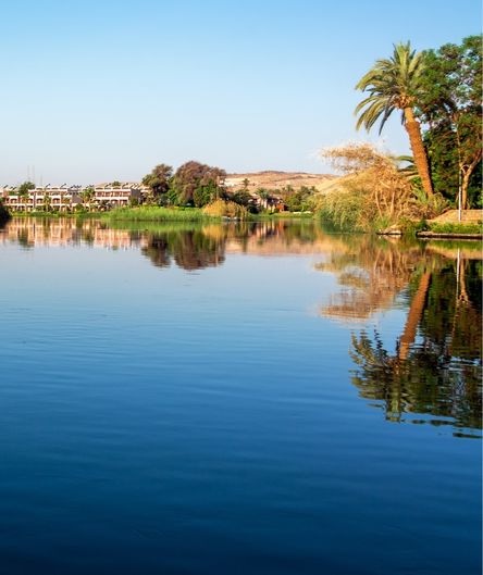 Ruhiges, blaues Wasser spiegelt Palmen und Gebäude am Ufer einer Oase. Im Hintergrund erstrecken sich grüne Bäume und flache Hügel. Ruhiges, blaues Wasser spiegelt Palmen und Gebäude am Ufer einer Oase. Im Hintergrund erstrecken sich grüne Bäume und flache Hügel.