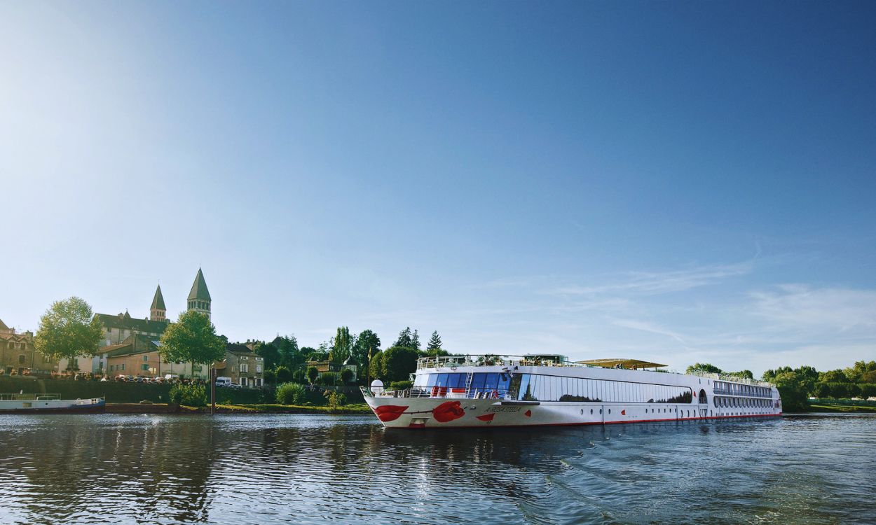 Weißes A-ROSA Flusskreuzfahrtschiff mit rotem Kussmund fährt auf einem Fluss an einer Stadt mit Kirchttürmen unter blauem Himmel vorbei. Weißes A-ROSA Flusskreuzfahrtschiff mit rotem Kussmund fährt auf einem Fluss an einer Stadt mit Kirchttürmen unter blauem Himmel vorbei.