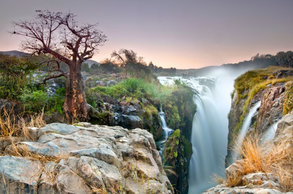Epupa-Wasserfälle in Namibia: Ein mächtiger Wasserfall stürzt in eine Schlucht, daneben ein markanter Affenbrotbaum (Baobab) auf Felsen im Abendlicht. Epupa-Wasserfälle in Namibia: Ein mächtiger Wasserfall stürzt in eine Schlucht, daneben ein markanter Affenbrotbaum (Baobab) auf Felsen im Abendlicht.