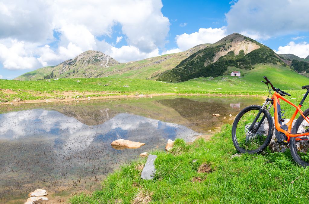 An einem Bergsee-Ufer steht ein orangefarbenes E-Mountainbike. Die Berge und der Wolkenhimmel spiegeln sich im Wasser. An einem Bergsee-Ufer steht ein orangefarbenes E-Mountainbike. Die Berge und der Wolkenhimmel spiegeln sich im Wasser.