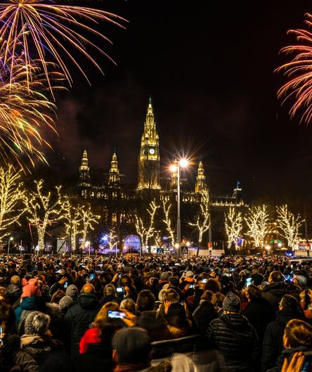 Ein großes Feuerwerk erhellt den Nachthimmel über dem beleuchteten Wiener Rathaus. Davor steht eine dichte Menschenmenge auf einem Platz. Ein großes Feuerwerk erhellt den Nachthimmel über dem beleuchteten Wiener Rathaus. Davor steht eine dichte Menschenmenge auf einem Platz.