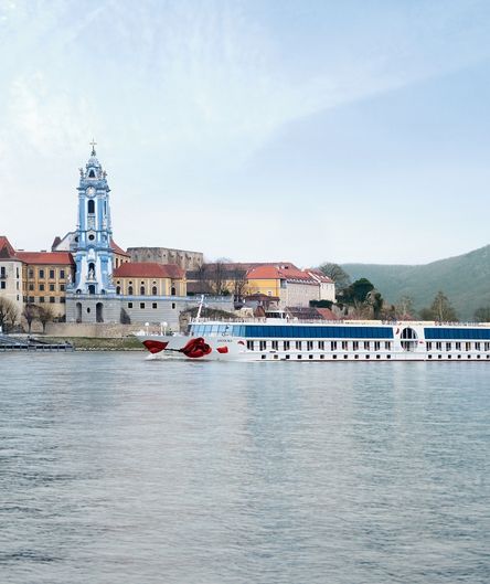 Ein Flusskreuzfahrtschiff der A-ROSA-Flotte passiert die Ortschaft Dürnstein in der Wachau mit dem blau-weißen Turm des Stiftes Dürnstein im Hintergrund.