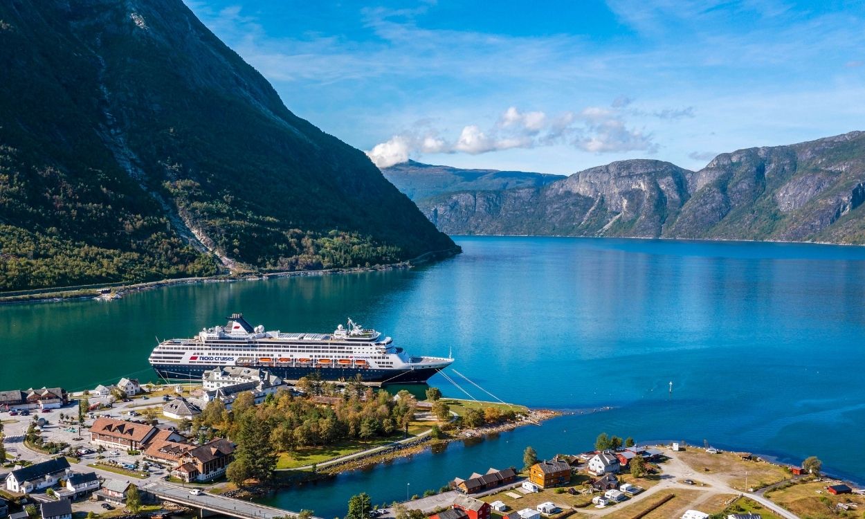 Kreuzfahrtschiff im tiefblauen Fjord von Geiranger, Norwegen, umgeben von hohen, bewaldeten Bergen und einem kleinen Dorf im Vordergrund.