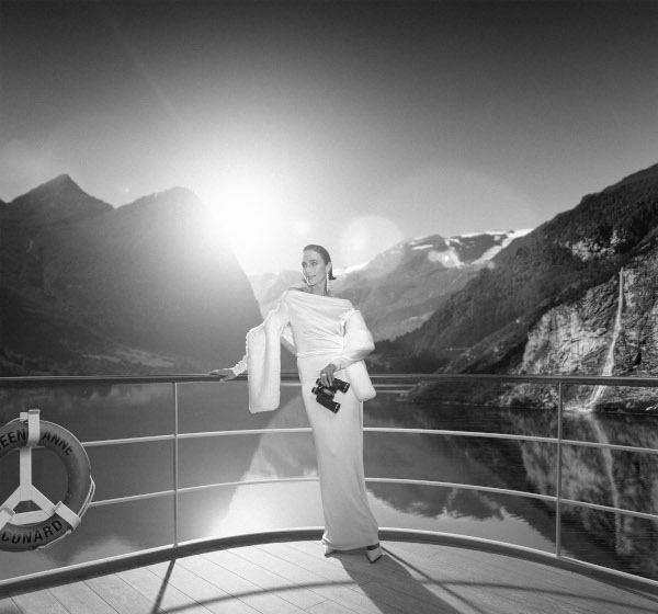 Schwarz-Weiß-Aufnahme: Frau im weißen Abendkleid mit Fernglas an einem Schiffsgeländer vor beeindruckender Kulisse aus Fjord und Bergen.